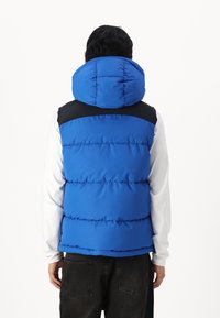 Veste rembourrée bleue avec une grande capuche, présentant un panneau supérieur noir. Texture douce, design matelassé et coupe décontractée. Portée sur une chemise à manches longues.