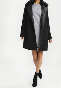 Schwarzer Wollmantel mit Pelzkragen, einer strukturierten Innenfütterung, Einzelknopfverschluss und geradem Schnitt. Über einem grauen Kleid getragen.