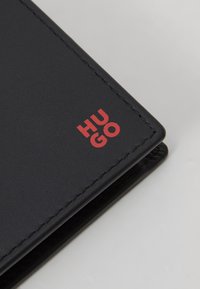 Portofel din piele neagră cu logo-ul roșu "HUGO". Prezintă linii curate, textură netedă și cusături pe margini. Formă compactă, rectangulară.