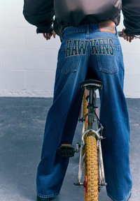 Svijetloplave široke traperice s natpisom "HAWKINS" na stražnjim džepovima, nošene dok sjedi na biciklu s žutim kotačem.