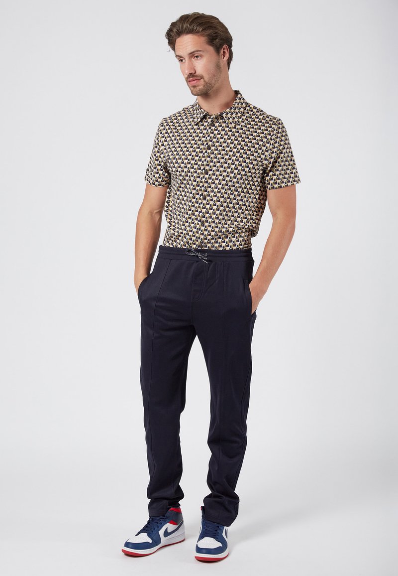 Koszula z krótkim rękawem w wielokolorowy geometryczny wzór, w połączeniu z granatowymi spodniami tapered i niebieskimi trampkami z czerwonymi akcentami.