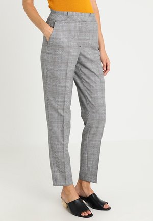 Mujer lleva pantalones de cuadros grises de talle alto con bolsillos laterales, combinados con mules de tacón abiertos en negro y un top mostaza.