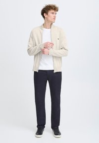 Suéter con cremallera beige claro sobre una camiseta blanca, combinado con pantalones azul marino oscuros y zapatillas negras. El suéter tiene puños acanalados y un logo.