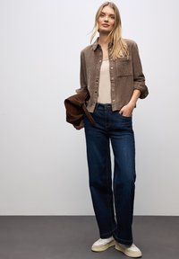 Camisa de pana marrón con botones, camiseta sin mangas beige, jeans de pierna ancha azul oscuro y zapatillas blancas. La modelo sostiene un bolso de hombro marrón.