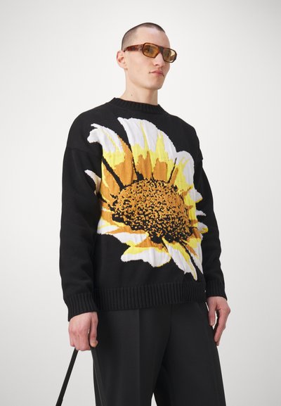 Schwarzer Strickpullover mit einem großen Sonnenblumenmuster in Gelb, Weiß und Braun. Gerippte Bündchen und Saum, getragen mit dunklen Hosen.