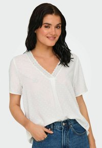 Vit kortärmad blus med V-ringning och spetsdetaljer, prydd med ett prickschema och lös passform, kombinerad med högmidjade blå jeans.