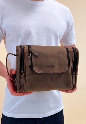 Man in white t-shirt holding a brown leather dual-zipper travel toiletry bag with handle on the side.