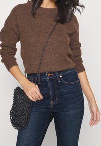 Persona vestida con un suéter de punto marrón y unos pantalones vaqueros de cintura alta de color azul oscuro, sosteniendo un bolso cruzado de red negro por la correa.