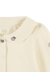 Cream-colored cotton shirt with a collar, featuring a pink embroidered flower and a round gray logo on the sleeve. Two snap buttons visible.