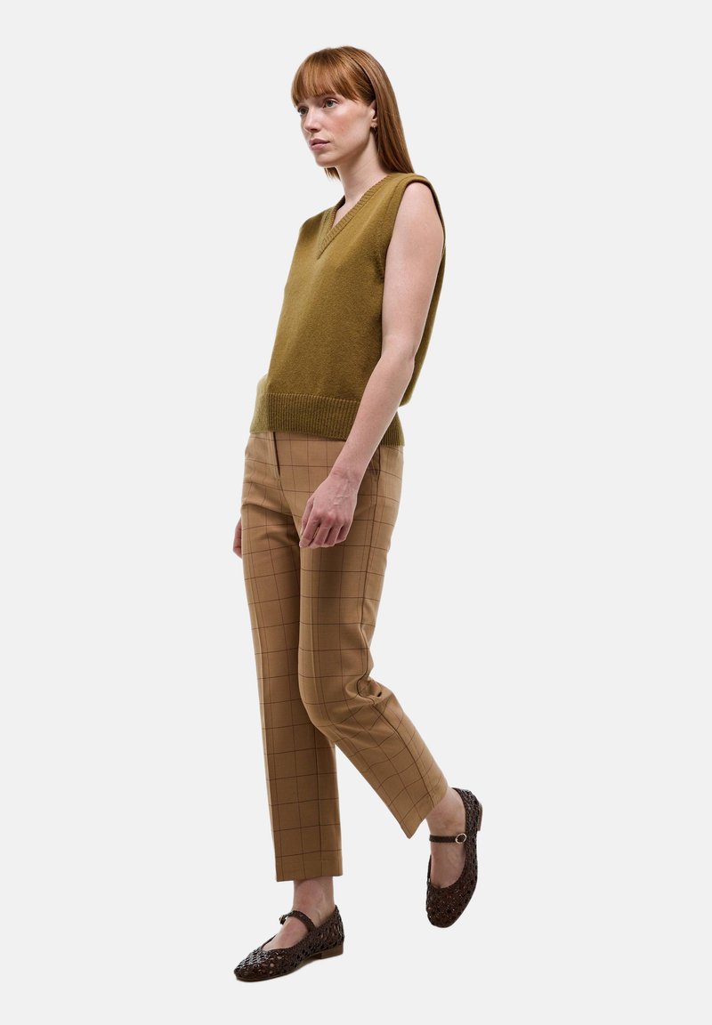 Pull sans manches vert olive avec un décolleté en V, assorti à un pantalon à carreaux marron taille haute et des ballerines marron à motifs.