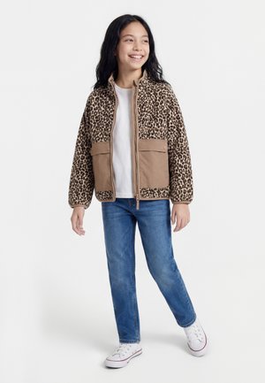 Ragazza sorridente, indossa una giacca in pile con stampa leopardata e tasche beige, t-shirt bianca, jeans blu e sneakers bianche, in piedi su sfondo bianco.