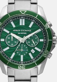 Zeleni številčnica ure s srebrnim nerjavečim jeklenim trakom. Ima tri podštevilke, debele oznake ur in prikaz datuma. Vodoodporna do 100 metrov.