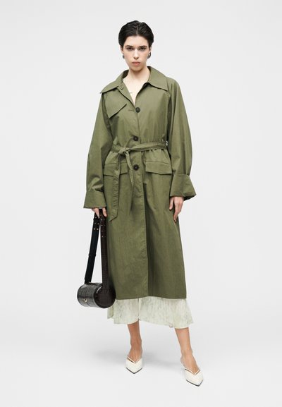 Πράσινο χακί παλτό τύπου trench με ζώνη για δέσιμο, μεγάλες μπροστινές τσέπες και φαρδύ γιακά· φορεμένο πάνω από ελαφρύ φόρεμα με λουλούδια και συνδυασμένο με λευκά παπούτσια μυτερά.