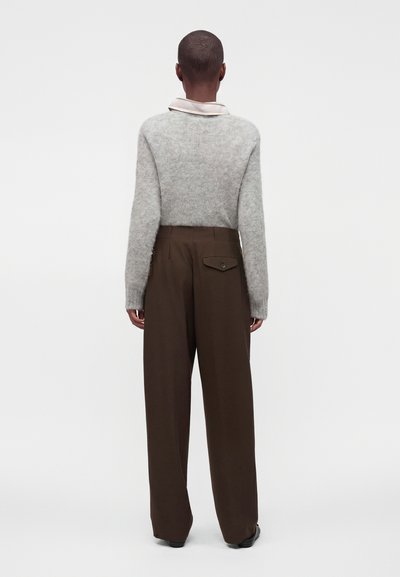 Personne debout dos à la caméra, portant un pull gris clair à manches longues et un pantalon marron ample taille haute. Fond uni.