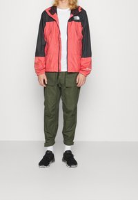 Červená a černá lehká bunda s kapucí, zipem a elastickými manžetami, doplněná zelenými cargo kalhotami a černými teniskami.