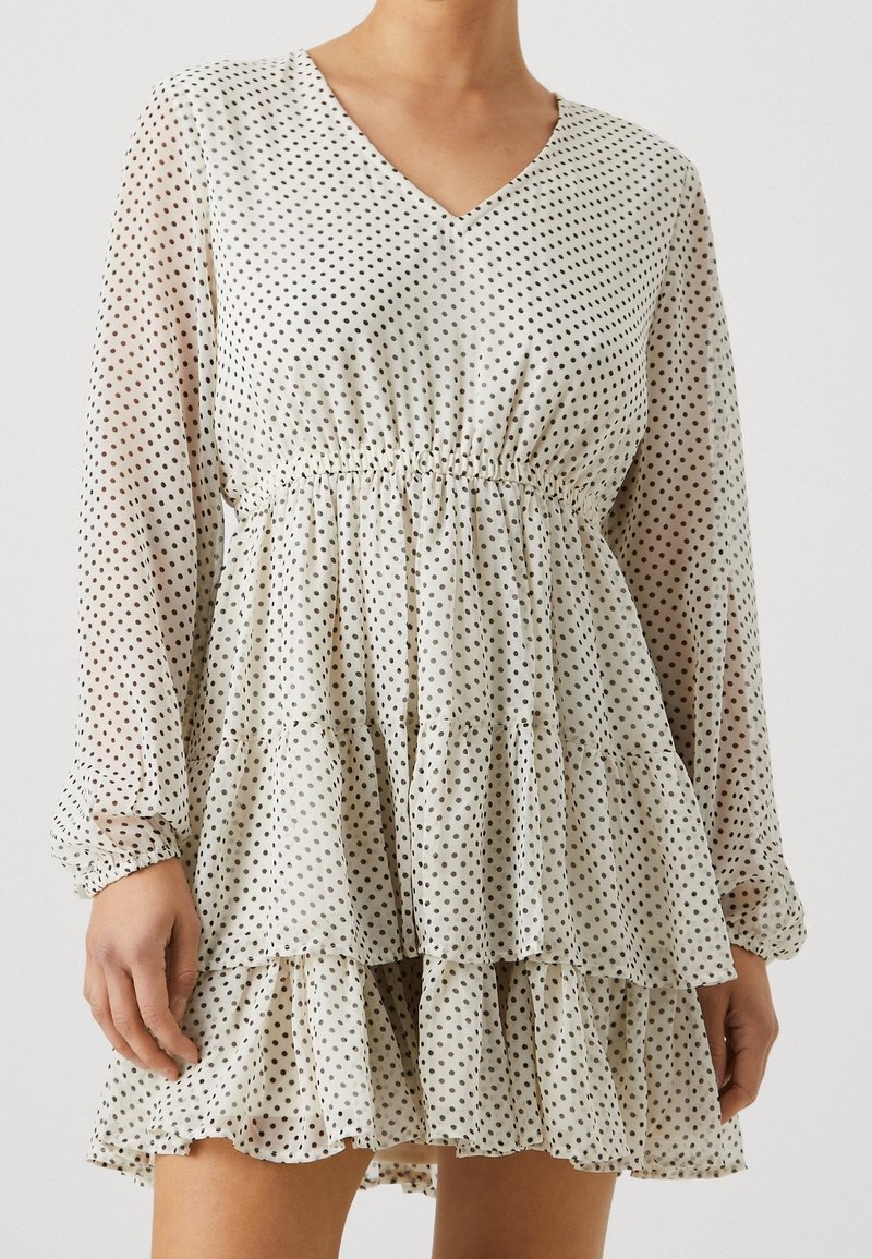 Femme portant une robe blanche à décolleté en V avec des pois noirs, des manches longues transparentes, une taille élastique et une jupe à volants étagée.