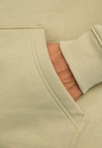 Világoszöld kapucnis pulóver anyaga, elülső zsebbel; bordázott mandzsetta részletek; látható varrás kiemelések; puha tapintású. A kéz részben a zsebben van.