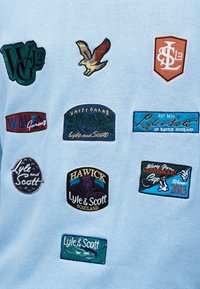 Ανοιχτό μπλε ύφασμα με εννέα πολύχρωμες κεντητές μπαλώματα, συμπεριλαμβανομένου ενός πουλιού που πετά και διάφορων λογότυπων και κειμένων της Lyle & Scott που αναφέρονται στη Σκωτία.