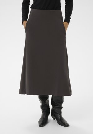 Eine Person trägt einen dunkelbraunen Midikleid mit den Händen in den Taschen, ein schwarzes Langarmoberteil und schwarze Lederstiefel mit Absatz vor einem weißen Hintergrund.