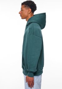 Grüne Stoffkapuzenjacke mit Kängurutasche, gerippten Bündchen und einer verstellbaren Kapuze, die ein minimalistisches, gesticktes Logo auf der Vorderseite hat.