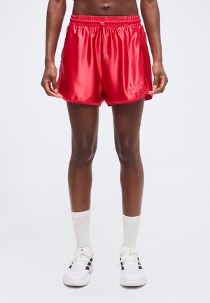 Henkilö, jolla on kiiltävät punaiset Adidas urheilushortsit, valkoiset vartta peittävät sukat ja valkoiset lenkkarit mustilla raidoilla, seisoo yksinkertaisen taustan edessä.