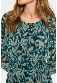 Žena v tyrkysových šatách s dlhými rukávmi, ktoré majú béžový a svetlomodrý listový kvetinový vzor a nariasený pás.