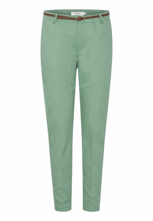 Nő áll, világosszürke, testhezálló rövid ujjú felsőt, menta zöld nadrágot barnás övvel és fehér tornacipőt visel sima háttér előtt.