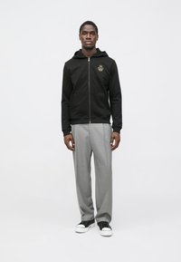 Sweatshirt preta com fecho éclair e capuz, apresentando um bolso frontal e um emblema dourado. Combinada com calças cinza claro e ténis pretos.