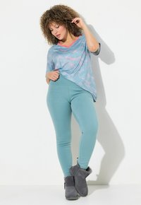 Donna con capelli ricci che indossa una blusa a fantasia blu chiaro, leggings turchesi e stivaletti alla caviglia foderati in pelliccia grigia, in posa su sfondo bianco.