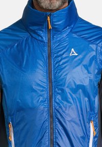 Veste zippée bleue avec col haut, arborant un logo blanc et des détails de fermeture éclair orange. Le matériau a une texture lisse et brillante.