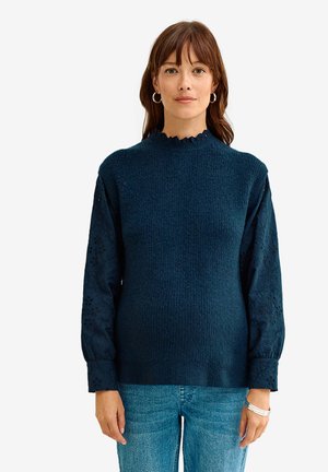 Frau trägt einen marineblauen, strukturierten Pullover mit gemusterten langen Ärmeln und hellblauen Jeans, blickt nach vorne vor einem weißen Hintergrund.