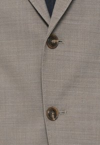 Μπεζ σακάκι με υφή από ύφασμα, διαθέτει καμπύλη πέτο και δύο καφέ κουμπιά στο μπροστινό μέρος. Ορατή λεπτομέρεια με υποτονικό σχέδιο πλέγματος.