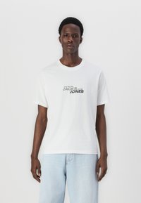 Camiseta de algodón blanca con el texto "JACK & JONES" en negro y gris. Corte casual con mangas cortas, combinada con jeans de mezclilla azul claro.
