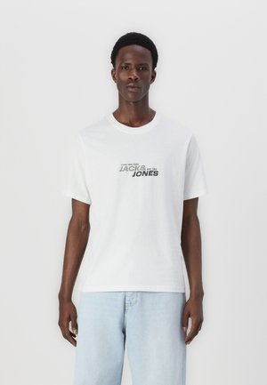 Camiseta de algodón blanca con el texto "JACK & JONES" en negro y gris. Corte casual con mangas cortas, combinada con jeans de mezclilla azul claro.