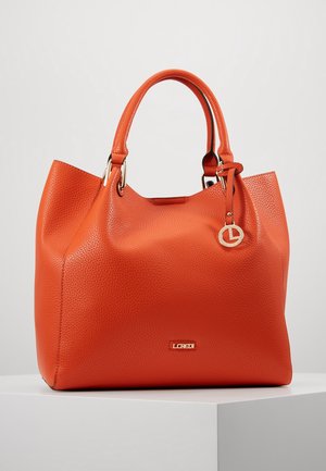 Sac à main en cuir orange texturé avec doubles poignées, plaque de logo dorée et pendentif circulaire doré sur un socle blanc sur fond uni.