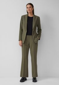 Un costume vert olive avec une veste ajustée et un pantalon droit, associé à un haut noir, porté avec des chaussures noires. Texture de tissu lisse.