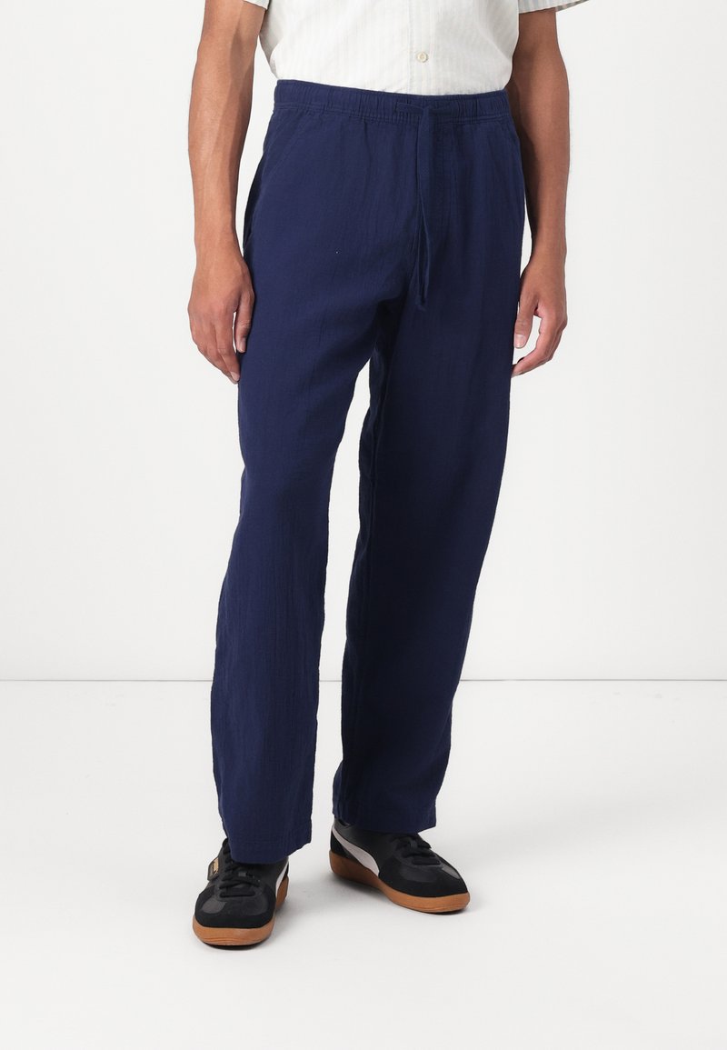 Pantalons en lin léger bleu marine avec une taille à cordon. Présentent une coupe ample et un tissu texturé pour le confort. Chaussures noires avec semelles en gomme.