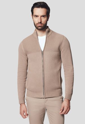 Beżowy sweter zapinany na zamek błyskawiczny o prążkowanej fakturze, z wysokim kołnierzem, bocznymi kieszeniami i dopasowanym krojem. Nosi się go na białej koszuli.