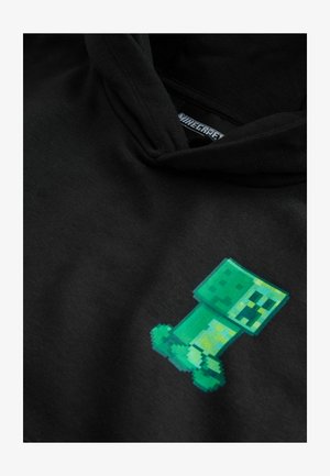 Juoda kapučiukė su pixeliniu žaliu Creeper personažu iš Minecraft ant priekinės pusės. Minkšta medžiaga, laisvas kirpimas, be matomų detalių.