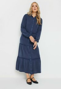 Marinblå knäppt klänning med långa ärmar, flerlagrad design och rynkade detaljer, tillverkad av mjukt tyg. Svarta platta skor bärs.