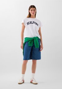 T-shirt bianca a maniche corte con la scritta "SCALPERS" in blu navy, abbinata a pantaloni corti in denim blu e un felpa verde legata in vita. Scarpe da ginnastica bianche.