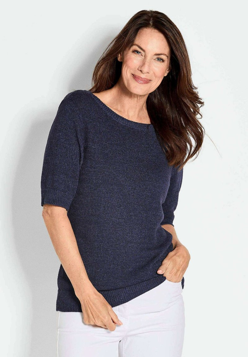 Marineblauer Strickpullover mit weitem Ausschnitt und kurzen Ärmeln. Der Stoff hat eine strukturierte Optik. Kombiniert mit weißen Hosen.