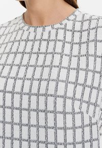 Top in maglia con scollatura rotonda, caratterizzato da un motivo a griglia nera su uno sfondo grigio chiaro. Texture morbida e vestibilità leggermente ampia.