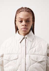 Jachetă crem matlasată cu un guler structurat, nasturi tip snap și două buzunare la piept. Prezentă logo-ul Nike pe partea stângă.