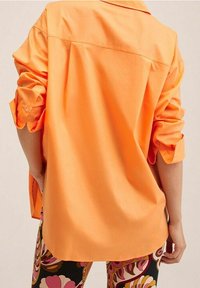 Chemise orange surdimensionnée en coton, avec un col classique et des manches retroussées, associée à des leggings à motifs aux couleurs vives.