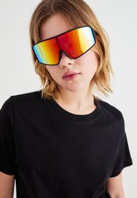 Sonnenbrille mit einem großen, kantigen Rahmen, reflektierenden orange- und blauen Gläsern sowie einem schwarzen Rand. Getragen über einem schwarzen T-Shirt.