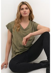 Oliivinvihreä V-pääntie t-paita, jossa on lyhyet hihat, löysä istuvuus ja hienovarainen textura, yhdistettynä mustiin legginsseihin ja nilkkureihin.