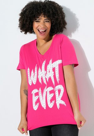 Hymyilevä nainen, jolla on kiharat hiukset, pukeutuneena kirkkaan vaaleanpunaiseen T-paitaan, jossa on valkoiset niititut kirjaimet "WHATEVER" yksivärisellä valkoisella taustalla.
