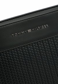 Sort Tommy Hilfiger buddeltaske har en vævet lædertekstur, glatte accenter og et metallisk logo på frontdesignet.