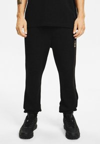 Mężczyzna w czarnych spodniach dresowych i czarnych trampkach, stojący z rękami swobodnie opartymi wzdłuż ciała na białym tle.